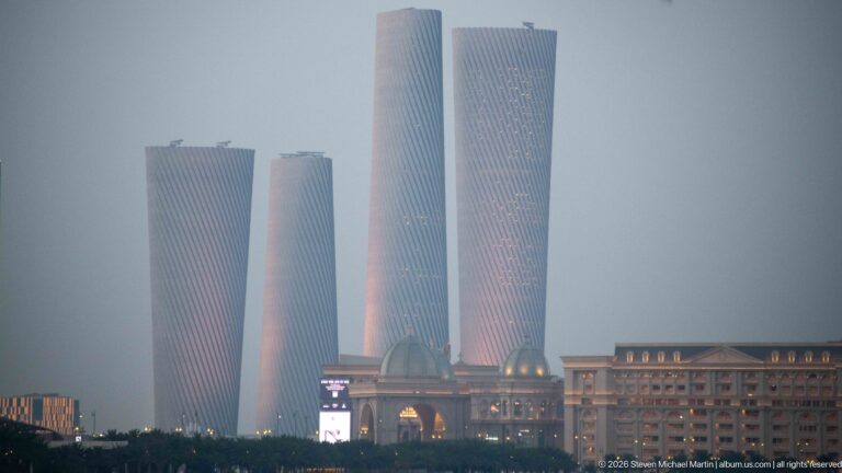 Four twisting Lusail Plaza Towers rise through haze above lower buildings in soft evening light.