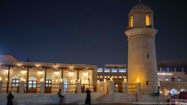 Tall cylindrical minaret stands beside a warmly lit mosque façade with arched windows under a dark blue sky.
