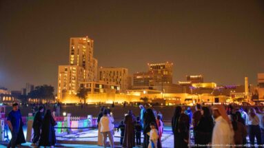 Crowds gather along a brightly lit promenade with warm-lit mid-rise buildings rising in the background.