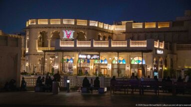 People gather outside an illuminated two-story arcaded building with balconies and restaurant fronts at night.