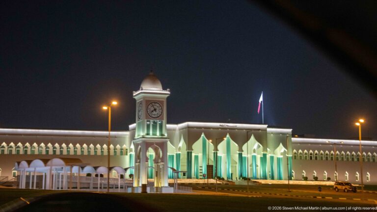 Illuminated white civic building with a central clock tower, pointed arches, and a Qatar flag at night.