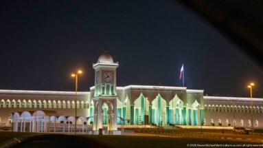 Illuminated white civic building with a central clock tower, pointed arches, and a Qatar flag at night.