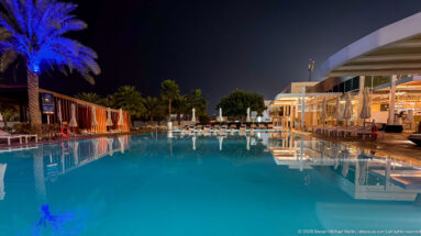 Large turquoise swimming pool extends toward palms, loungers, and a brightly lit covered terrace at night.