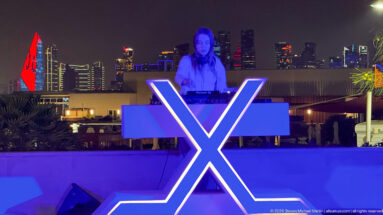 DJ stands behind a glowing X-shaped console, with Doha's illuminated skyline visible in the distance.