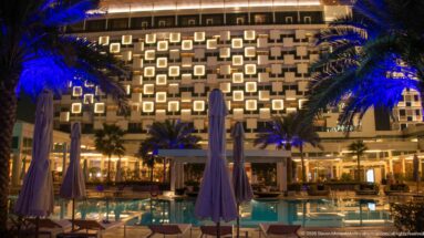 Closed parasols stand beside a swimming pool facing an illuminated hotel façade and blue-lit palms.