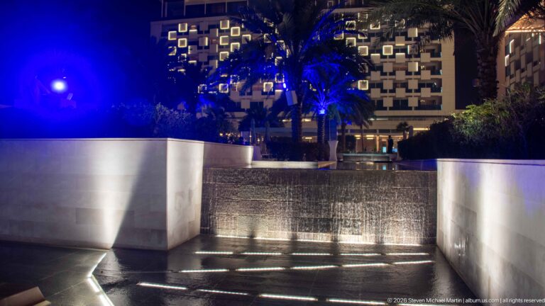 Water cascades over a stepped edge between pale walls, with recessed lights and blue-lit palms behind.