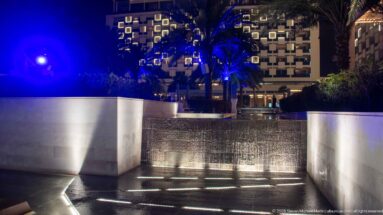 Water cascades over a stepped edge between pale walls, with recessed lights and blue-lit palms behind.