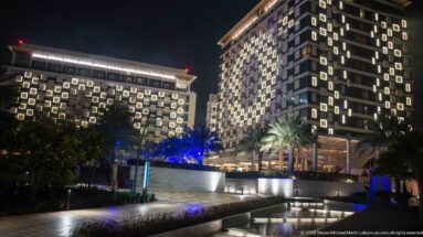 Two modern towers with glowing square window surrounds rise above palms, planted beds, and low walls at night.