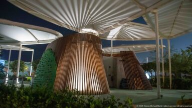 White radial canopy roofs fan out above tapered timber-slat columns beside planted landscaping at night.