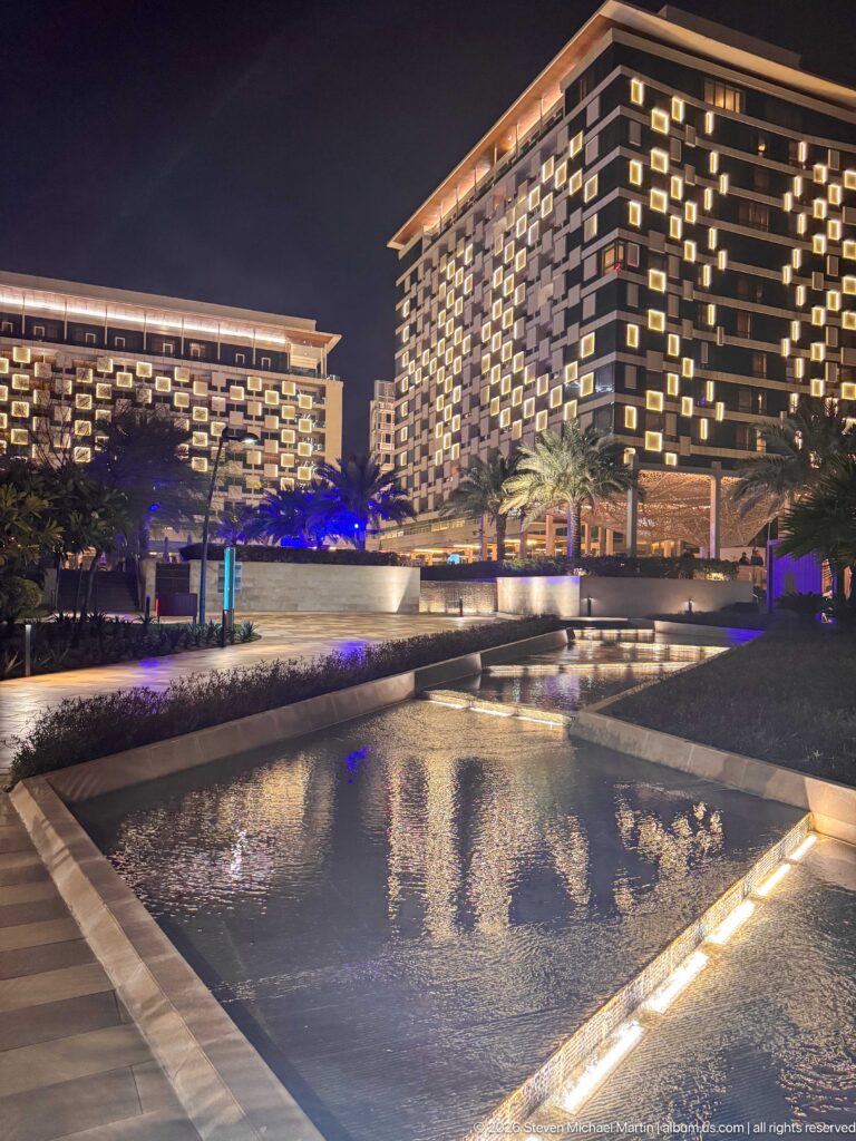 Two illuminated modern hotel blocks rise behind reflecting pools, palms, and low landscaped terraces at night.
