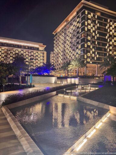 Two illuminated modern hotel blocks rise behind reflecting pools, palms, and low landscaped terraces at night.