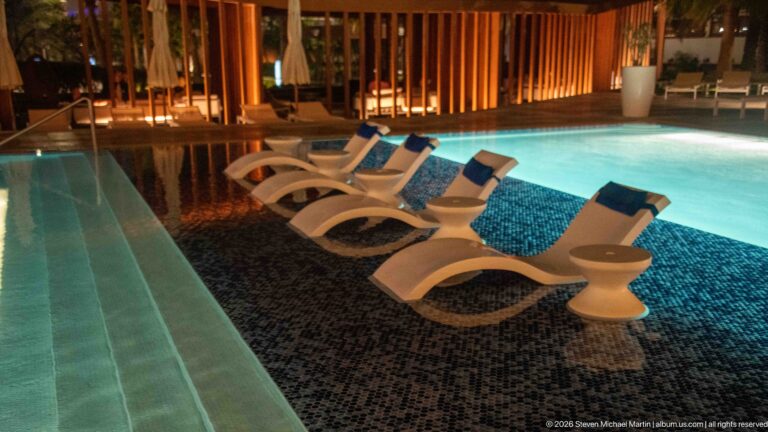 White sculptural loungers partly submerged in a turquoise-lit pool beside warm wooden screening at night.