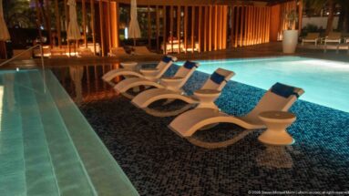 White sculptural loungers partly submerged in a turquoise-lit pool beside warm wooden screening at night.