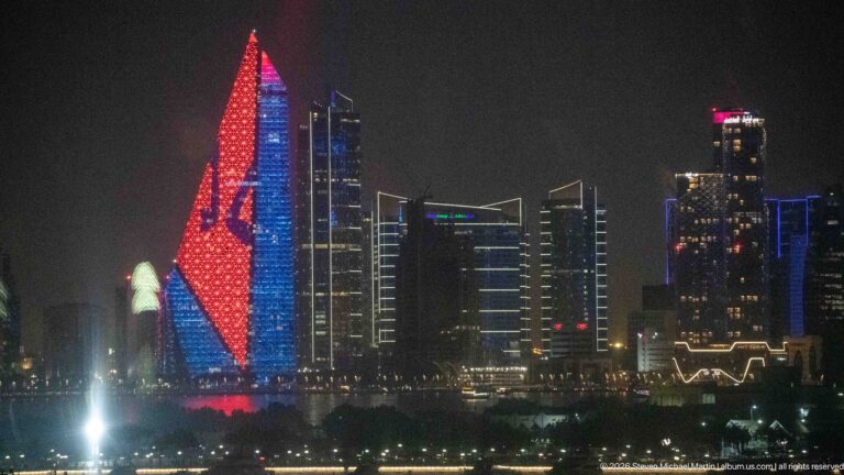 Tall tapered tower illuminated in red and blue at night, with darker high-rise buildings surrounding it.