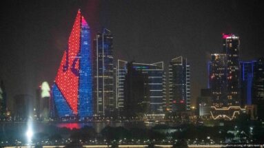 Tall tapered tower illuminated in red and blue at night, with darker high-rise buildings surrounding it.
