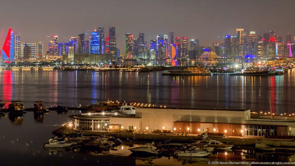 Wide nighttime view across Doha Bay toward the West Bay skyline, with marina buildings and boats in the foreground.