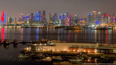 Wide nighttime view across Doha Bay toward the West Bay skyline, with marina buildings and boats in the foreground.