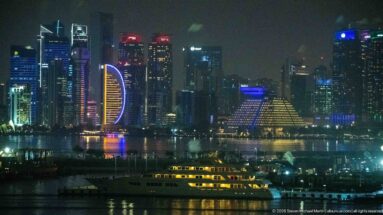 Illuminated passenger vessel on Doha Bay with the brightly lit West Bay skyline and tower reflections behind it.