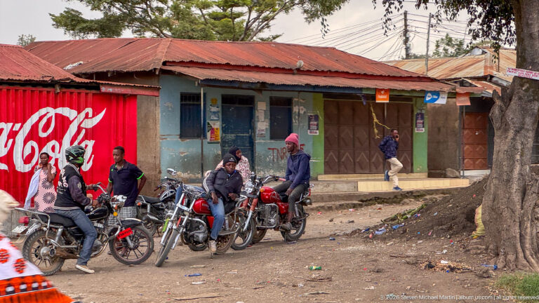 Group of motorcyclists gathered beside a large tree and roadside shops, with a red Coca-Cola panel behind them.