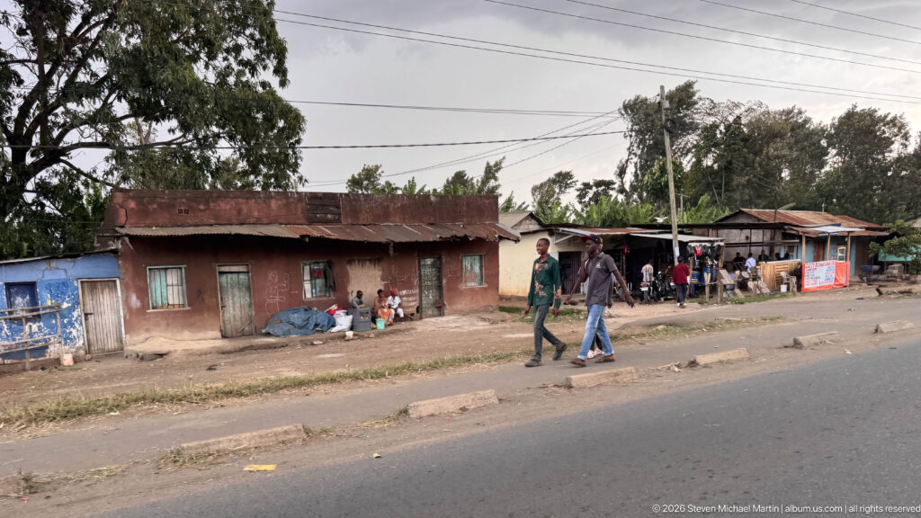 Two pedestrians walking beside weathered single-story buildings and small roadside shops along a paved Arusha street.