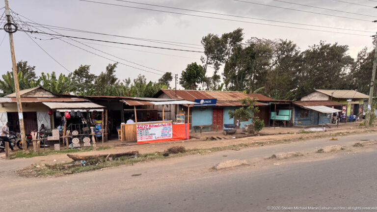 Row of small roadside shops with corrugated roofs, open storefronts, signs, trees, and utility wires beside an Arusha road.