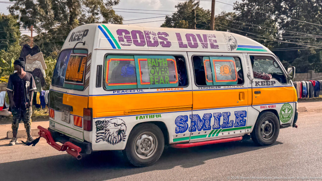 Colorfully painted minibus marked GOD'S LOVE, FAITH OVER FEAR, FAITHFUL, and SMILE beside roadside clothing displays.