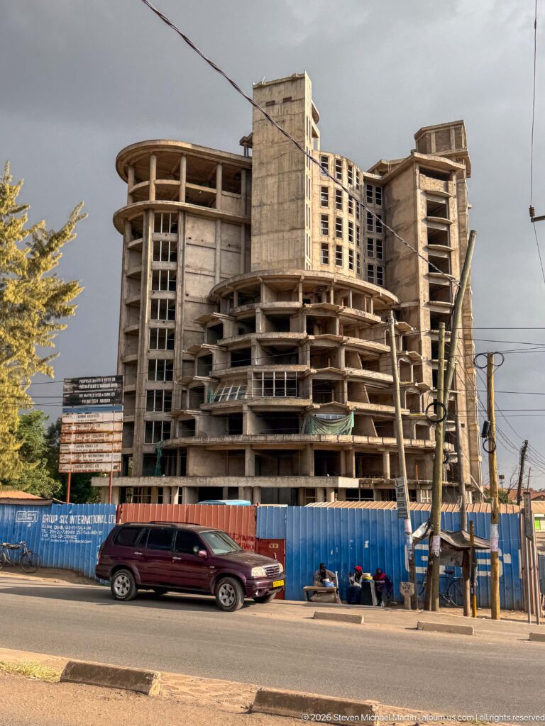 Large unfinished concrete building with exposed floor slabs, columns, open bays, and blue construction fencing beside an Arusha road.