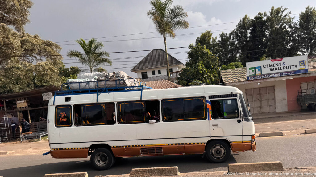 White passenger minibus with seated riders and luggage tied to a roof rack traveling along an Arusha street.