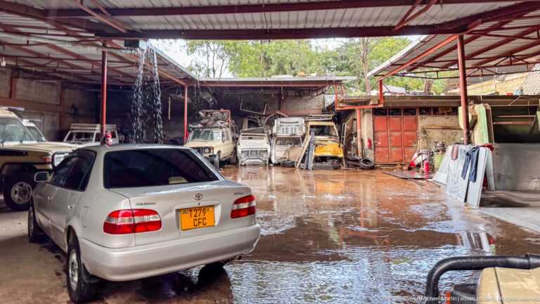Covered auto workshop with a silver sedan, older vehicles, body panels, tools, and wet concrete flooring.