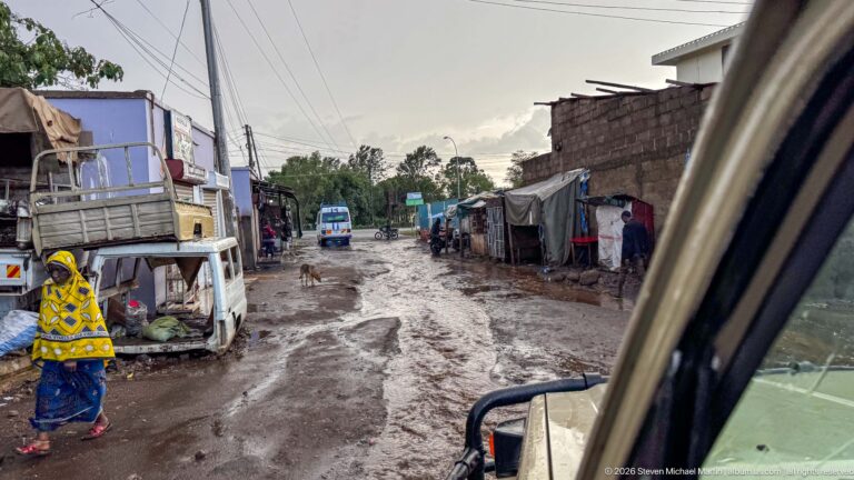 Muddy rutted street with standing water, small roadside structures, trucks, pedestrians, and a dog, viewed from a vehicle.