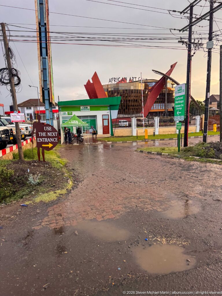 Wet Arusha roadside with puddles, utility lines, signs, and the red African Art Gallery building in the background.