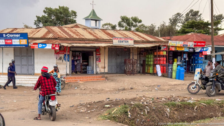 Roadside retail center with shops, motorcycles and pedestrians, with a small cross-topped church tower rising behind the roofs.