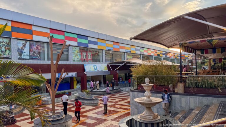 Elevated view of a patterned commercial courtyard with a tiered fountain, pedestrians, terraces, and a brightly paneled façade.