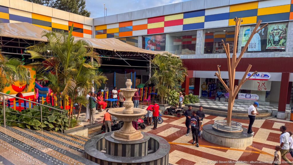 Open-air shopping courtyard with a tiered fountain, patterned paving, palms, visitors, and a multicolored building façade.