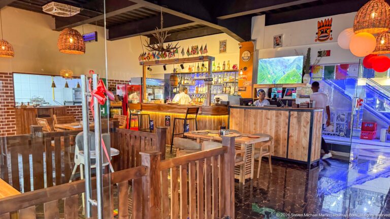 Wood-furnished bar and dining area with woven pendant lamps, colorful decorations, a stocked counter, staff, and patrons.
