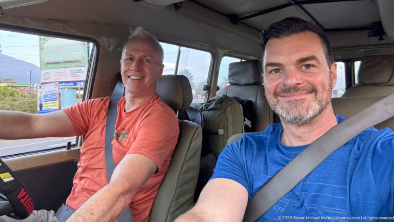 Two smiling travelers seated in the front of a large vehicle, with roadside buildings visible through the windows.