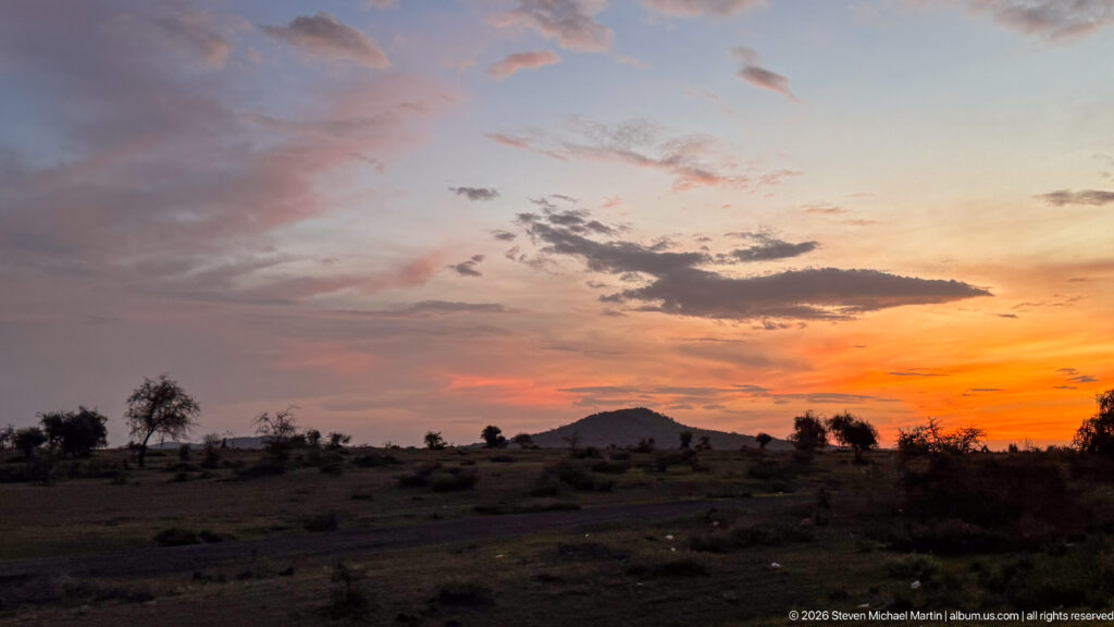 Orange sunset beneath layered clouds over a distant low mountain and scattered trees near Arusha.