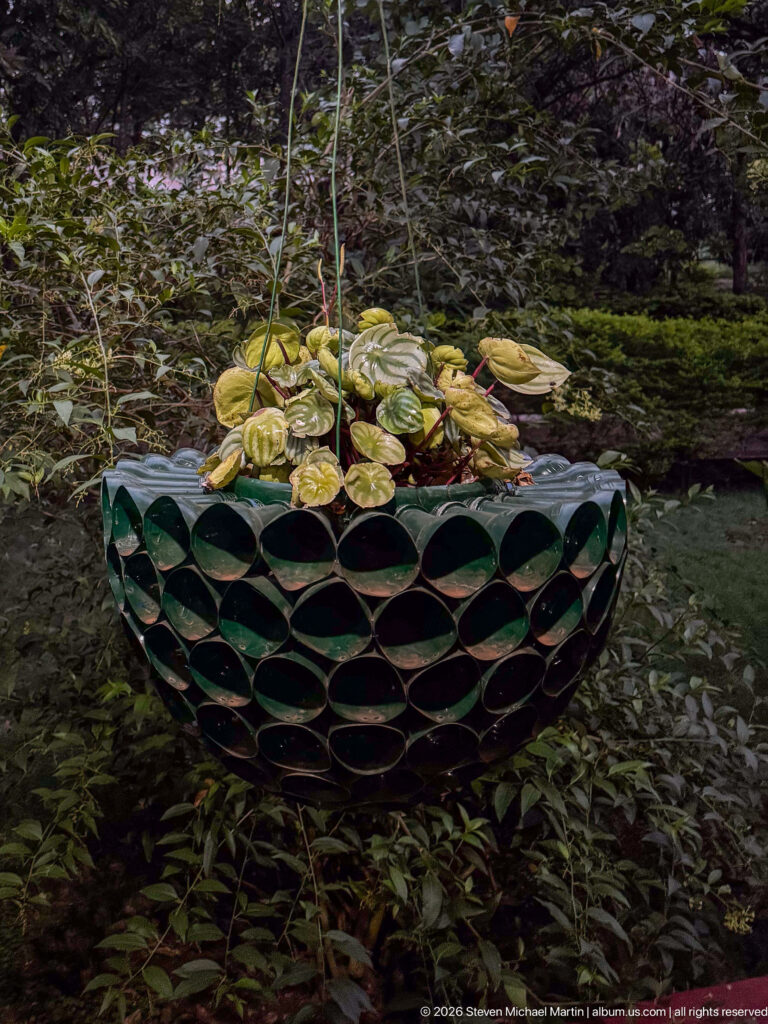 Round hanging planter made from dark cylindrical pieces, filled with variegated plants and suspended among garden foliage.