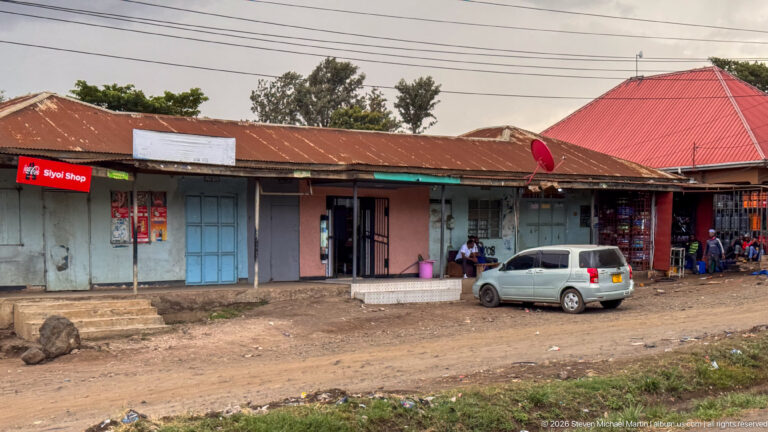 Weathered roadside shop row with painted doors, open retail bays, seated people, and a silver car parked in front.