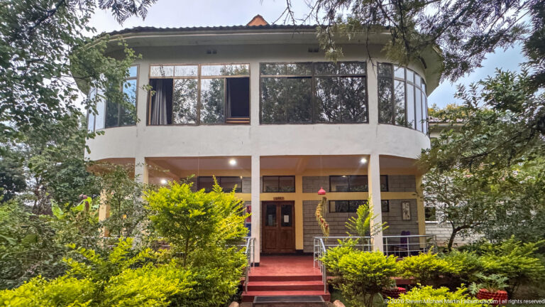 Two-story garden lodge with a broad windowed upper level, entrance steps, shrubs, and mature trees.