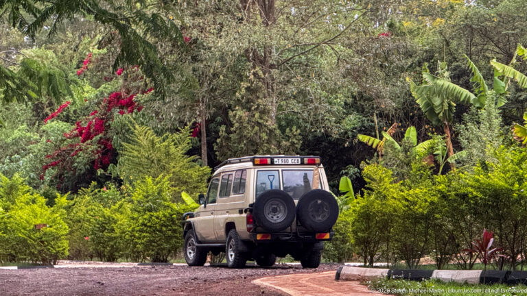 Four-wheel-drive vehicle with two rear spare tires parked on a gravel drive amid dense tropical garden vegetation.