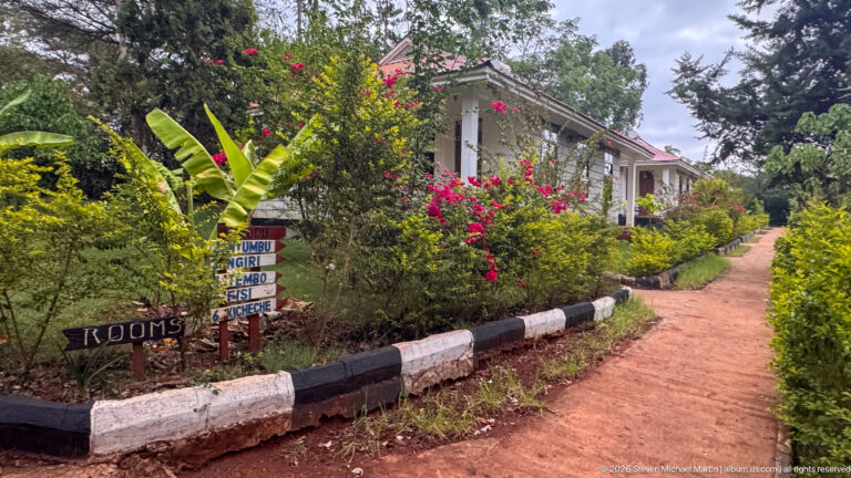 Red-earth path through a lush garden toward a light-colored guest building, with hand-painted room signs beside the walkway.