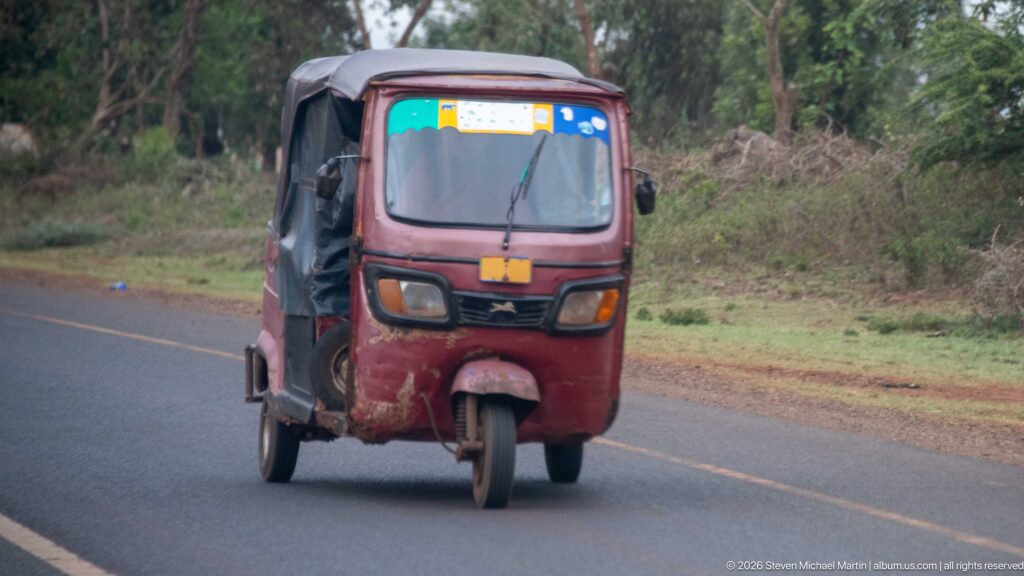 Red enclosed three-wheeled motor taxi driving toward the camera on a paved, tree-lined road.