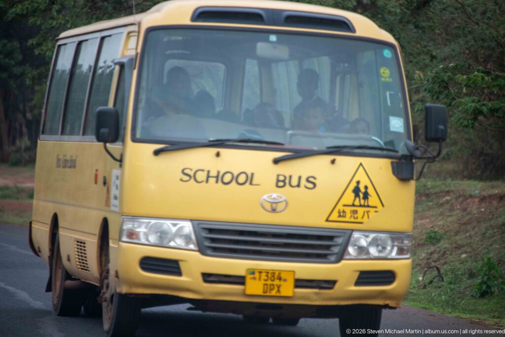 Front view of a yellow Toyota school bus driving toward the camera on a tree-lined road in Arusha.