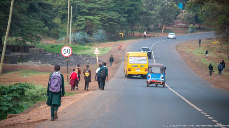 Curving Arusha road with pedestrians, a yellow school bus, blue three-wheeled taxi, cars, and a 50 km/h sign.
