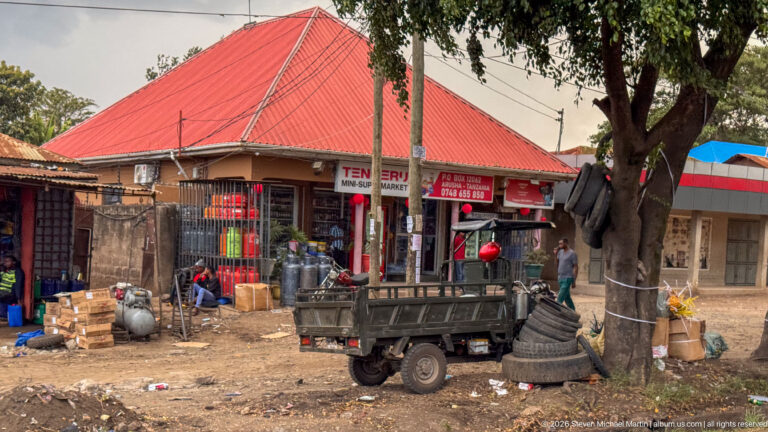 Roadside hardware shop displaying metal cylinders, tubing and stacked goods beside a small flatbed three-wheeler and large tree.