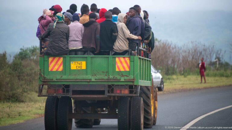 Rear view of a green open-bed truck crowded with standing passengers traveling along a paved road.