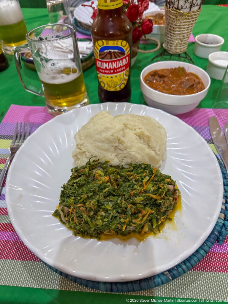 White plate with a mound of ugali beside cooked leafy greens, with Kilimanjaro beer and sauce on the table.
