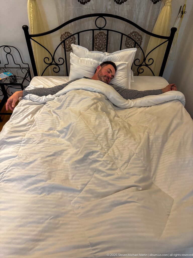 Guest resting under white bedding in a hotel room with a black wrought-iron headboard and warm bedside lighting.