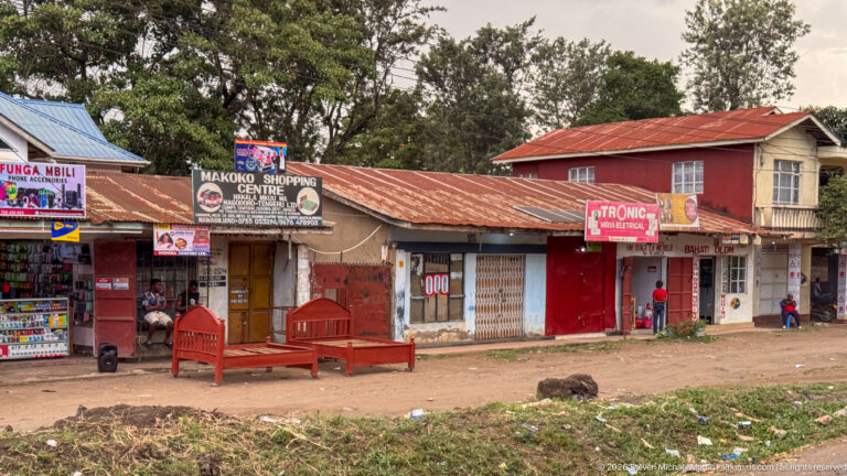 Makoko Shopping Centre and neighboring small storefronts beneath rusted corrugated roofs, with signs and a wooden bench outside.